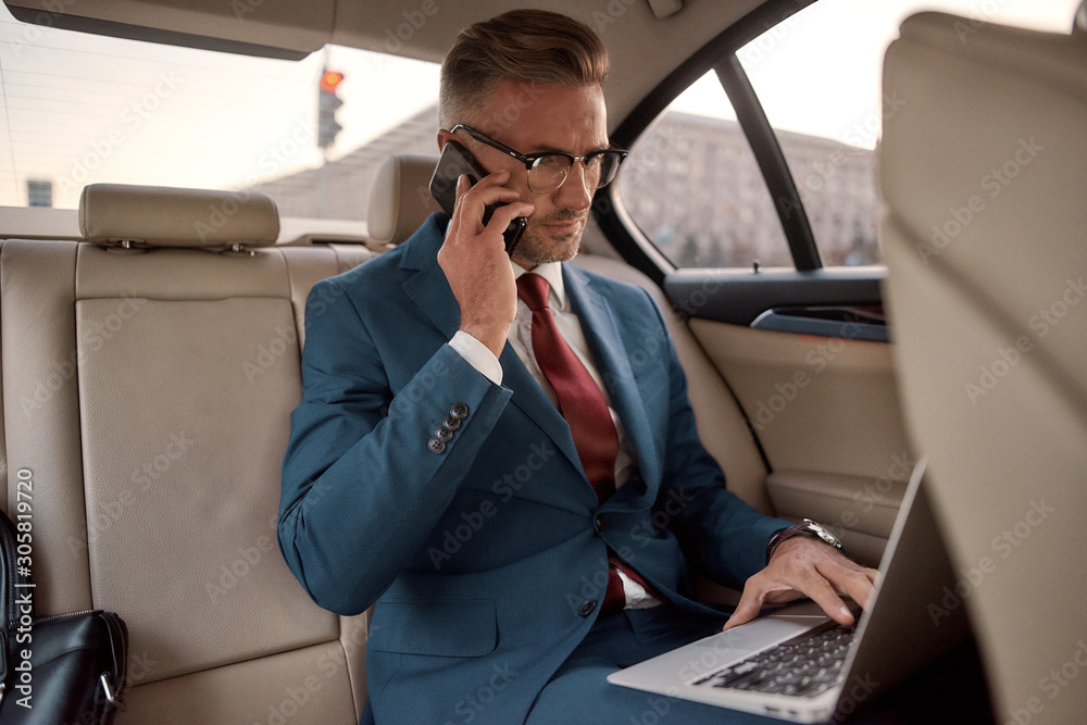Discussing project details. Confident and stylish mature businessman in full suit working on his laptop and talking on the phone with business partner while sitting in the car