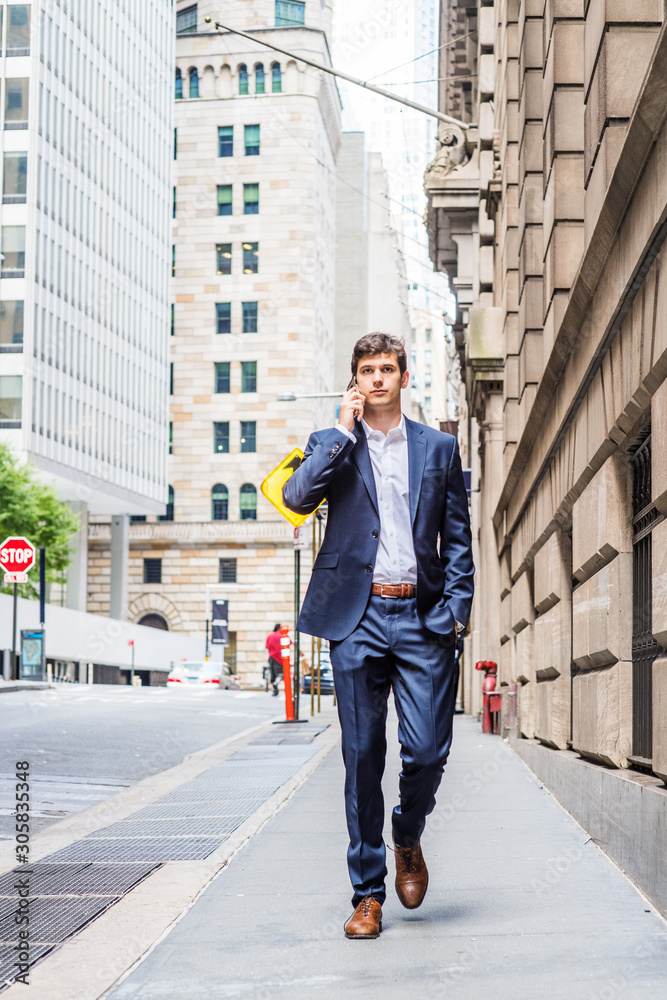Modern daily life. Young handsome man traveling in New York City, wearing blue suit, white shirt, brown leather shoes, hand in pocket, walking on street with high buildings, talking on cell phone..