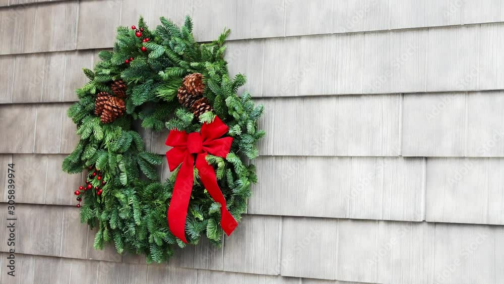 Christmas wreath with red bow is placed by hands of a woman in red shirt who hangs it on a home exterior wall. Christmas wreath with red bow is placed by hands of a woman in red shirt who hangs it on a home exterior wall.