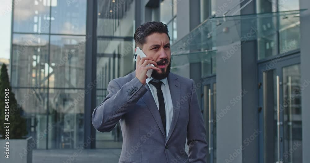 Busy serious Caucasian businessman speaking on the mobile phone with angry face as deciding some business issues and finishing his talk at the street while walking the city. Busy serious Caucasian businessman speaking on the mobile phone with angry face as deciding some business issues and finishing his talk at the street while walking the city.