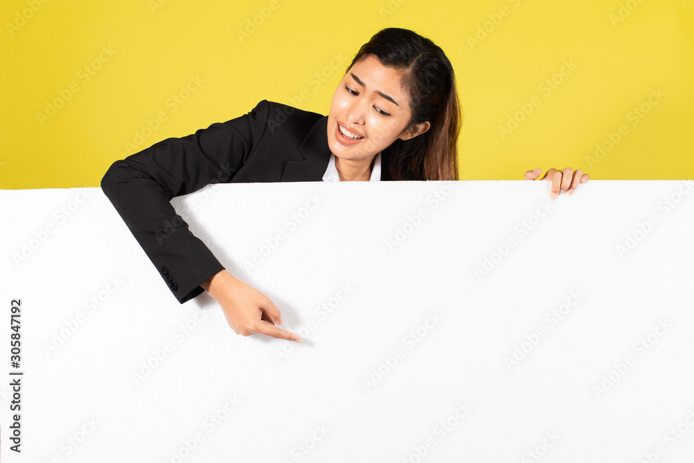 Young Asian woman in formal wear smiling and pointing at white blank banner for advertisement campaign against yellow background Young Asian woman in formal wear smiling and pointing at white blank banner for advertisement campaign against yellow background