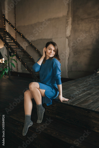 pretty brunette girl in a blue knitted dress without print, in shiny lurex socks sitting in a dark wood room against a background of light bokeh