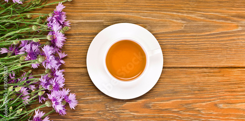 Black tea in a cup with pink cornflowers on a wooden background. Top view. Copy space.