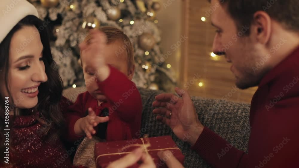 Close Up of Happy Caucasian Parents Helping Their Baby Son Open Present Box Wearing in Winter Sweaters sitting in Cosy Living Room on Grey Sofa near Christmas Tree Smiling Close Up of Happy Caucasian Parents Helping Their Baby Son Open Present Box Wearing in Winter Sweaters sitting in Cosy Living Room on Grey Sofa near Christmas Tree Smiling