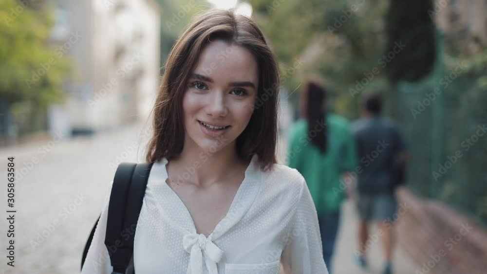 Portrait of Pretty Brunette Student Girl with a Bag Looking to Camera with a Smile. Young Woman Standing at Old City Street at the Background. Close Up shot. Portrait of Pretty Brunette Student Girl with a Bag Looking to Camera with a Smile. Young Woman Standing at Old City Street at the Background. Close Up shot.