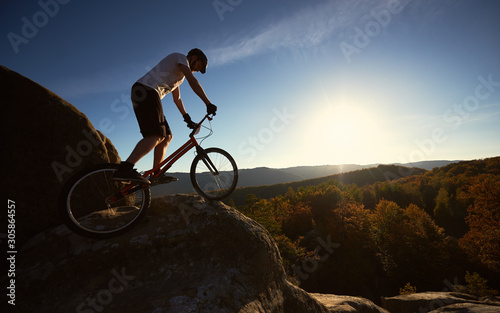 Young male cyclist balancing on trial bicycle, rider making acrobatic stunt on top of big boulder on summer sunny evening, blue sky and sunset on background.