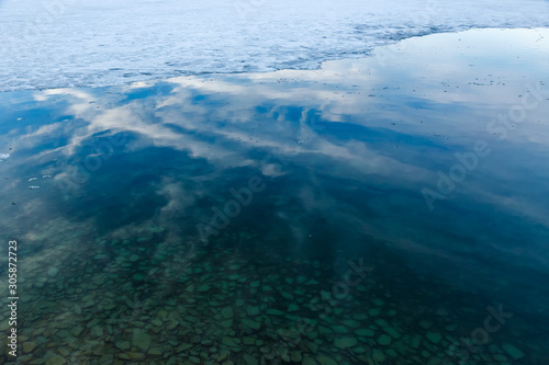 Border of melting ice and water of Baikal lake with pebbles under transparent water and reflection of sky with clouds