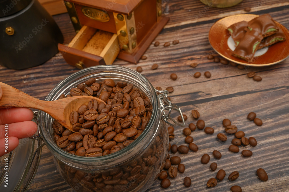 The girl's hand takes a wooden spoon of coffee beans from a glass jar, a coffee grinder, pieces of chocolate on a wooden background. Close up.