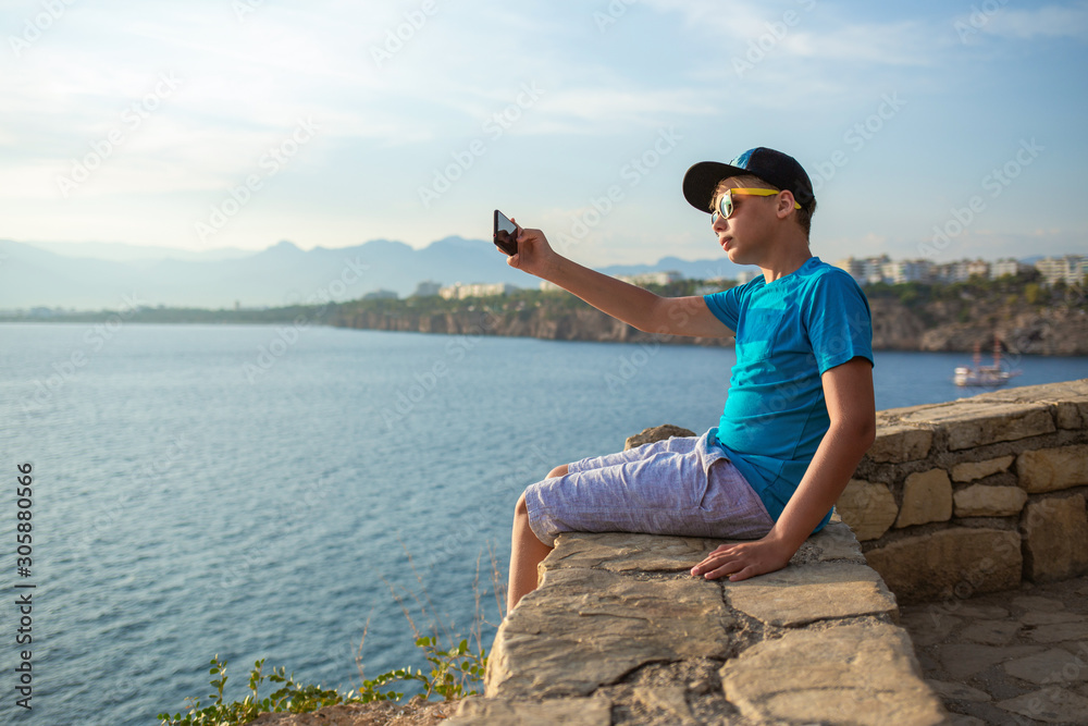 Young kid taking selfie using modern mobile smartphone posing outdoor at sunny scenic seascape. Happy travel and modern technology concept. Horiozntal color photography. Young kid taking selfie using modern mobile smartphone posing outdoor at sunny scenic seascape. Happy travel and modern technology concept. Horiozntal color photography.