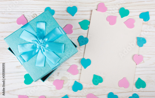 Blue gift box, paper hearts of pastel colors and a blank greeting card on the white wooden background (flat lay)