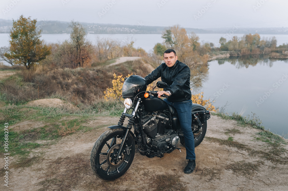Obraz premium Stylish man in a black leather jacket sits on an expensive vintage motorcycle. Portrait of a brutal biker, gathered on a journey against the backdrop of nature in cloudy weather. Photography, concept.