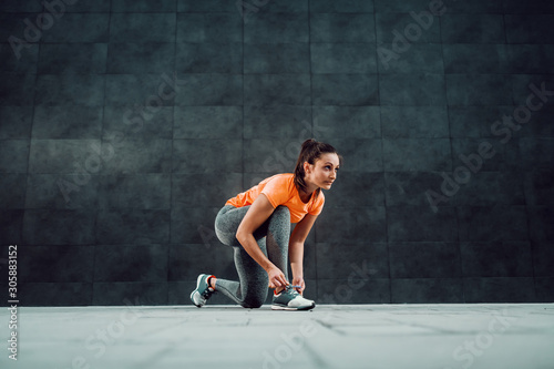 Fit attractive caucasian brunette in sportswear and with ponytail kneeling and tying shoelace before running.