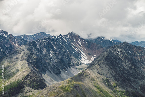 Fototapeta Naklejka Na Ścianę i Meble -  Atmospheric alpine landscape with big mountain ridge and snowbound peak in low clouds. Cloudy sky over huge mountain range. Giant snowy craggy rocks and rocky valley. Majestic scenery on high altitude