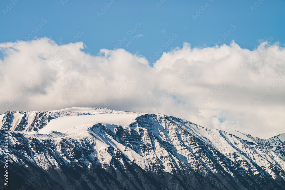 Atmospheric alpine landscape to snowy mountain ridge in sunny day. Snow shines in day light on mountain peak. Beautiful shiny snowy top. Wonderful scenery to low clouds above range covered with snow.