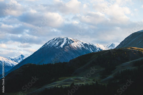 Atmospheric alpine landscape to snowy mountain ridge and forest hills in sunset. Snow shines in golden light on mountain peak. Wonderful scenery with beautiful shiny snowy top. Evening cloudy sky.