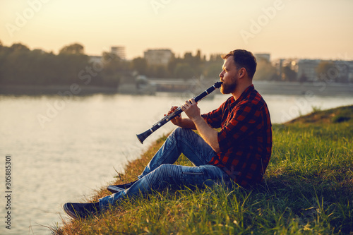 Canvas Print Side view of handsome caucasian bearded blond man in plaid shirt sitting on cliff and playing clarinet