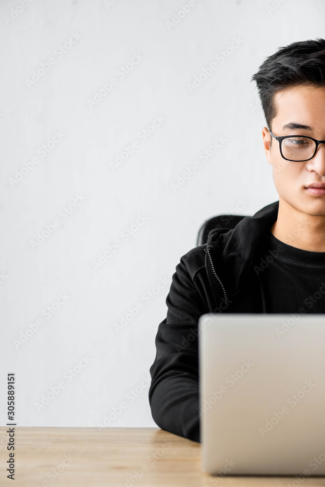 cropped view of asian hacker in glasses sitting at table and using laptop cropped view of asian hacker in glasses sitting at table and using laptop