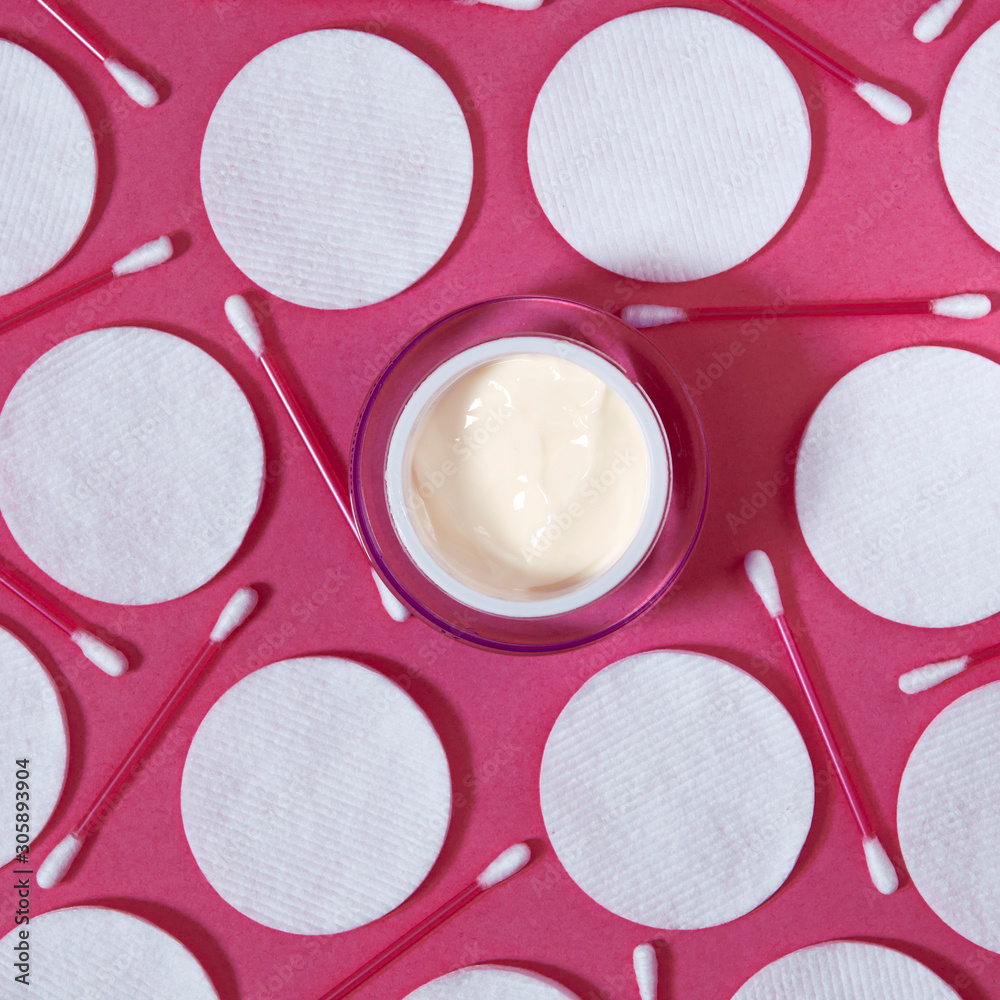 Hygiene products: white round cotton pads and plastic sticks for cleaning the ears lie on a pink background next to face cream Hygiene products: white round cotton pads and plastic sticks for cleaning the ears lie on a pink background next to face cream