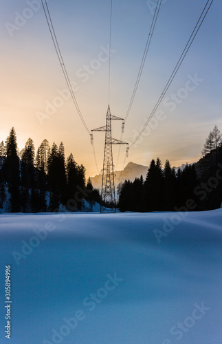 Power pole and power lines at sunset in a valley in winter. Fresh snow in foreground, forest and mountains in background with yellow hue
