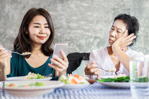 Asian woman using mobile phone ignoring mother while eating dinner together , social addiction concept 
