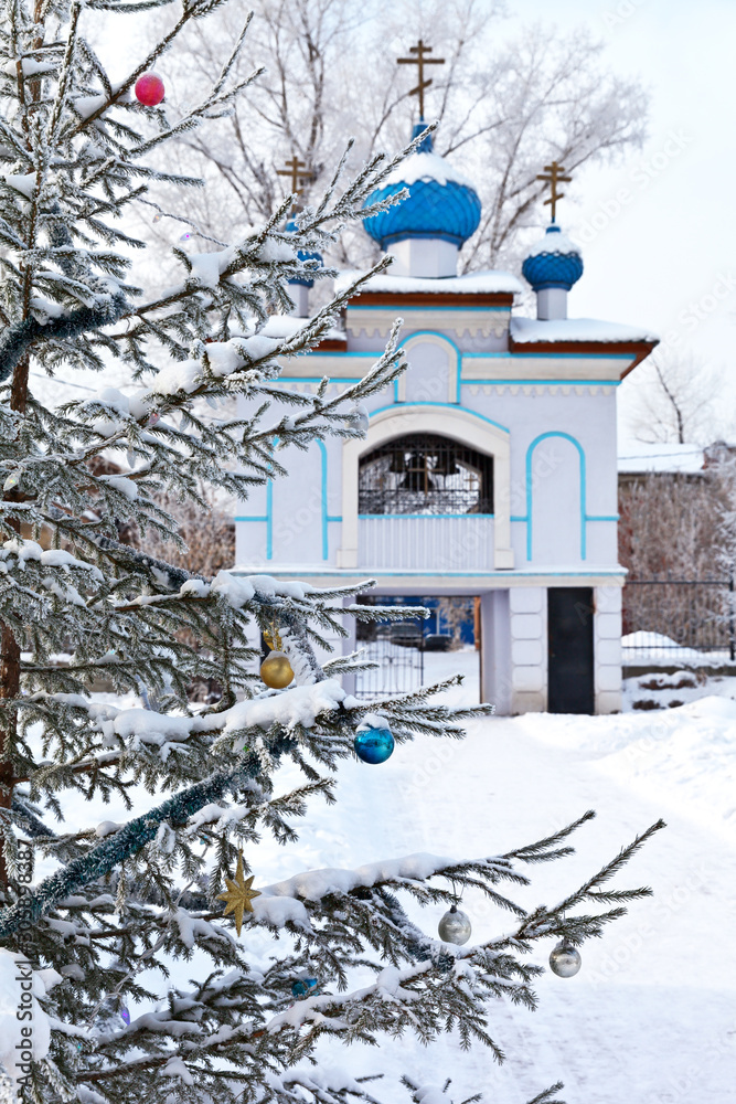 Fototapeta premium Irkutsk for Christmas holidays. Snow-covered Christmas tree with toys in the courtyard of the Church of the Assumption of the Mother of God on the background of a beautiful bell tower with blue domes
