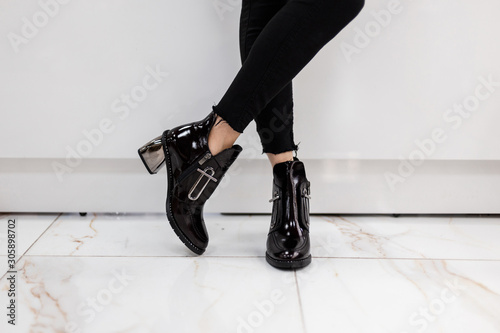 Fashionable young woman in stylish black jeans in trendy patent leather shoes with silver heel stands near a white wall in a shopping center. Girl chooses autumn boots. Shopping. Close-up.