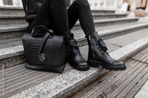 Young woman sits on stone steps outdoors in jeans in black fashion boots with a stylish leather handbag. Сlose-up of female legs in fashionable shoes with a trendy bag. Autumn winter collection.