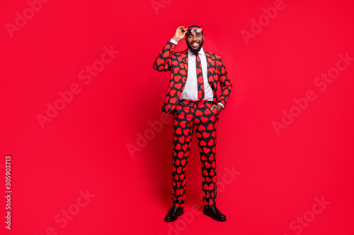 Full body photo of funky dark skin man playing amour cupid character role self-confident wear sun specs hearts pattern suit shirt necktie tie boots outfit isolated red color background