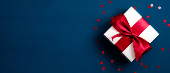 White gift box with red ribbon bow on blue background with confetti. Christmas present, valentine day surprise, birthday concept. Flat lay, top view.