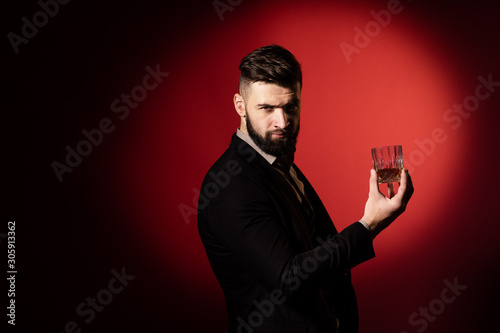 Handsome bearded man in suit with stylish beard holding a glass of whiskey and looks into the camera, on red background. Copy space © Olha