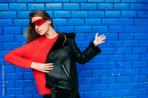 Fashion portrait of young long haired brunnete woman in black sheepskin coat, red sportive sunglasses posing over blue brick wall outdoors. photo