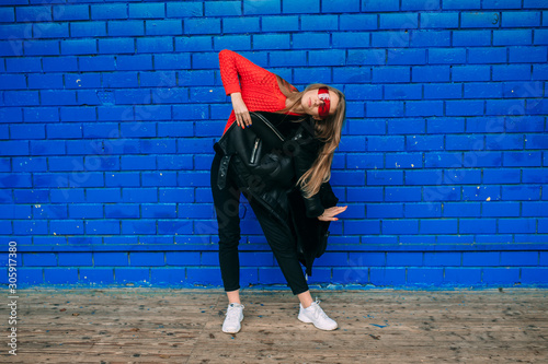 Fashion portrait of young long haired brunnete woman in black sheepskin coat, red sportive sunglasses posing over blue brick wall outdoors. photo
