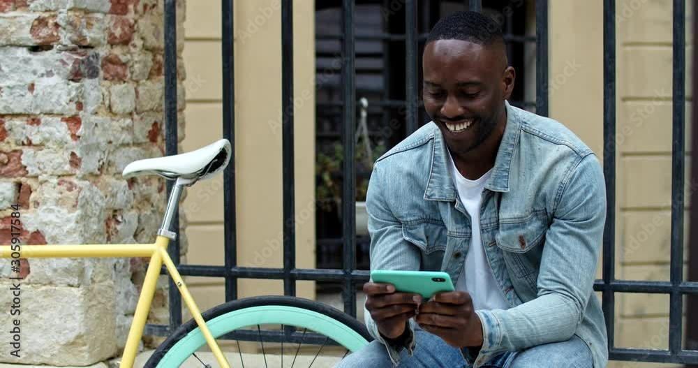 Cheerful African American Young Guy Using his Smartphone for Watching Videos, Looking Excited and Laughing while Sitting near Stylish Bike with Old Metal Fance at the Background.
