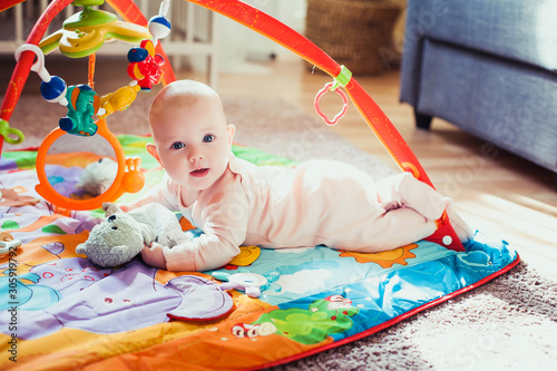 4 months old baby girl lying on colorful play mat on the floor. Activity carpet for kids. Early development at home.
