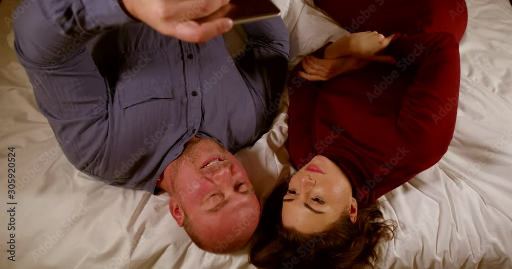The view from the top. An adult couple in the evening resting in bed watching video on a smartphone. A bald, unshaven man in a shirt and a woman in a dress to lie down. The view from the top. An adult couple in the evening resting in bed watching video on a smartphone. A bald, unshaven man in a shirt and a woman in a dress to lie down.