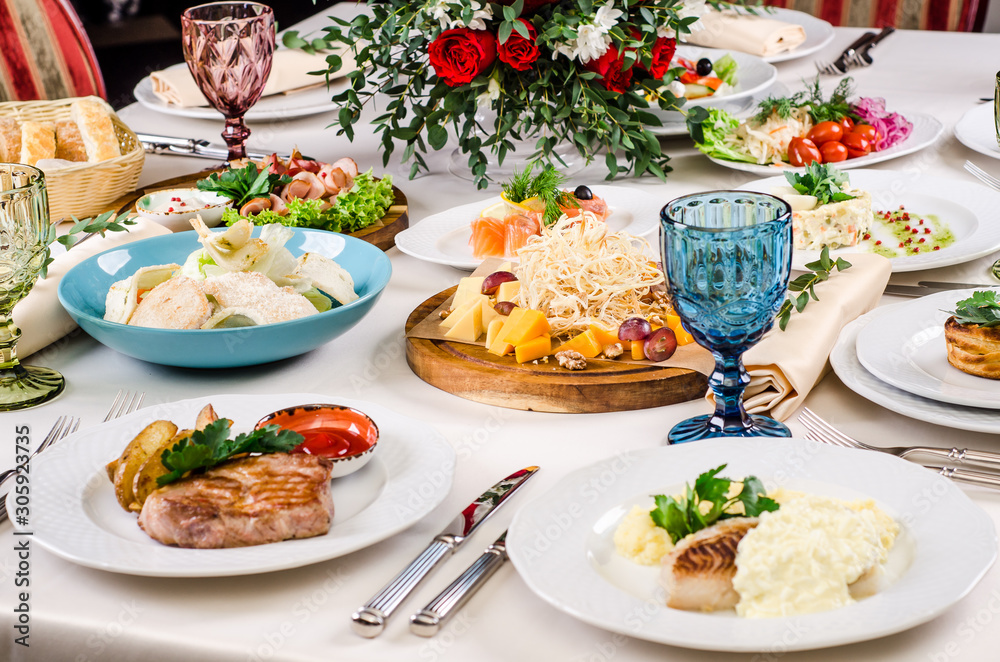 Grilled pork with potatoes and fried cod white fish with snack, salads, cutlery, wine glasses on banquet restaurant table background. European food in a restaurant setting.