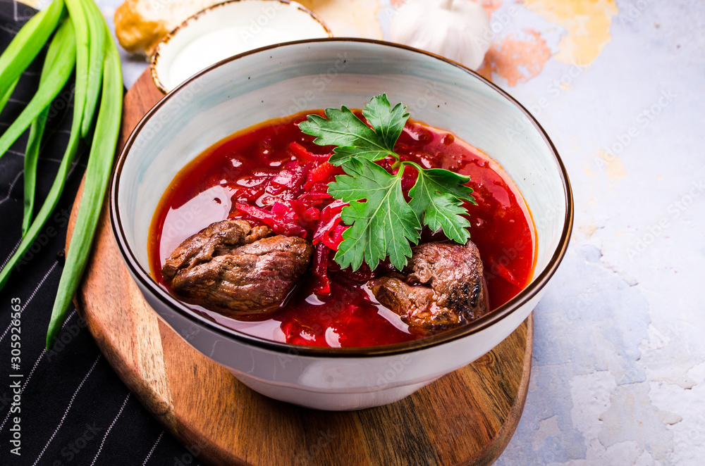 Traditional Ukrainian Russian Borscht with bacon, hot soup with beets, cabbage and carrots in a bowl on a wooden board. Served with green onions, sour cream, rye bread, garlic, greenery. Traditional Ukrainian Russian Borscht with bacon, hot soup with beets, cabbage and carrots in a bowl on a wooden board. Served with green onions, sour cream, rye bread, garlic, greenery.