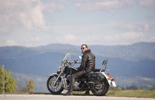 Handsome bearded biker with long hair in black leather jacket and sunglasses sitting on modern motorcycle on roadside  on blurred copy space background of dark woody hills and light foggy sky.