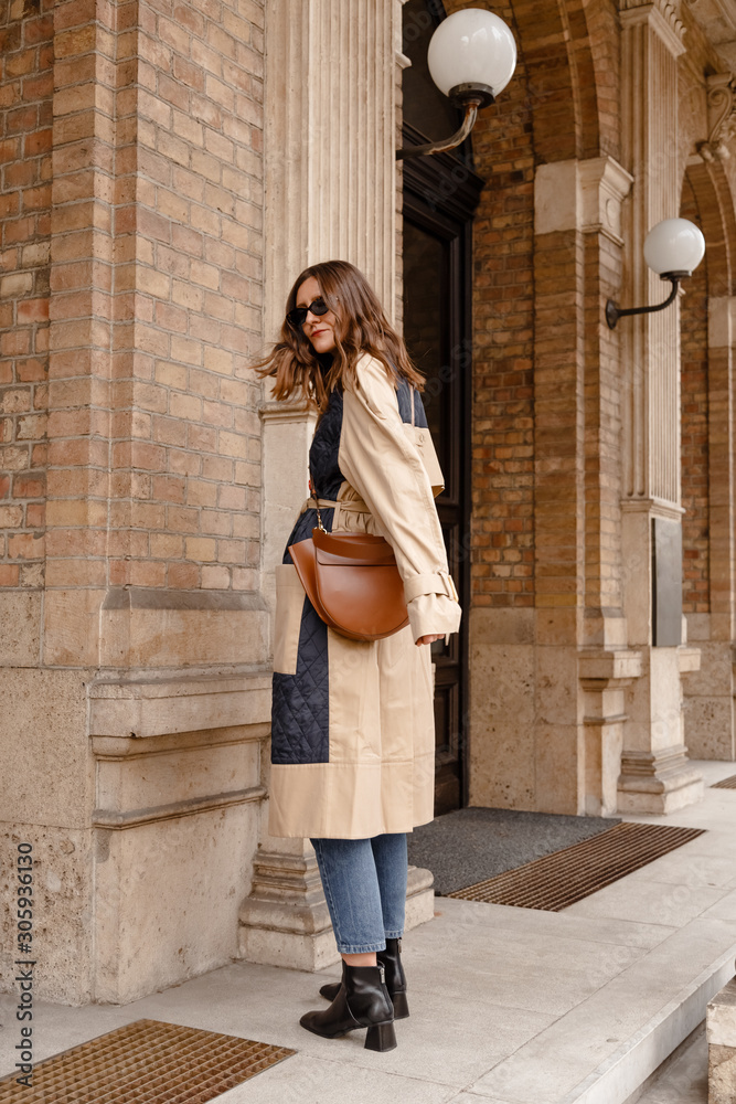 street style portrait of an attractive woman wearing a beige trench coat,  denim jeans, black ankle boots and brown leather bag, crossing the street.  fashion outfit perfect for autumn fall winter Stock