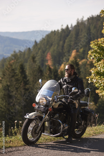 Fototapeta Naklejka Na Ścianę i Meble -  Handsome bearded biker in black leather jacket and sunglasses resting on cruiser motorcycle on country roadside, on blurred background of foggy green hills covered with dense spruce forest.