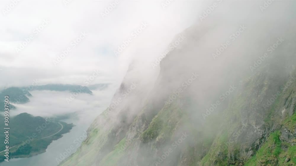 Scenic 4K wide drone shot with forward reveal motion of clouds and fog climbing steep, majestic and green mountain range and dark cliffs in overcast summer daylight, in Lofoten archipelago Norway. Scenic 4K wide drone shot with forward reveal motion of clouds and fog climbing steep, majestic and green mountain range and dark cliffs in overcast summer daylight, in Lofoten archipelago Norway.