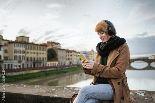 Young woman with red hair listens to music with headphones and uses the smartphone sitting on a bridge in Florence - Millennial is having fun and takes some time off