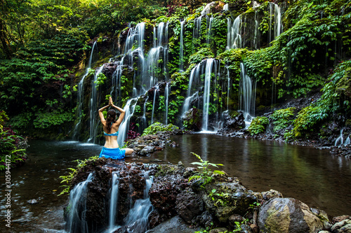 Woman sitting on the rock, practicing yoga. Young woman raising arms with namaste mudra near waterfall. Banyu Wana Amertha waterfall, Bali. View from back. photo