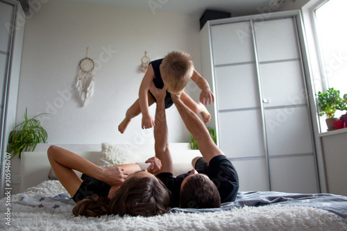 Happy family at home bedroom have fun  with kid on the bed together. People wearing black t-shirt,  shorts and baby bodysuit.  The dad holdinh the child on arms, the baby flying photo