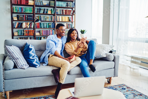 Cheerful African American couple having fun together at cozy living room sitting on sofa with smartphone, young hipster guys taking selfie on mobile phone camera recreating on leisure at home interior
