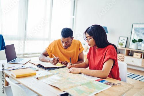 Thoughtful african american male and female colleagues sitting at desktop with map making route for exploring in future journey, serious hipster guys making planning of journey wiring notes indoors.
