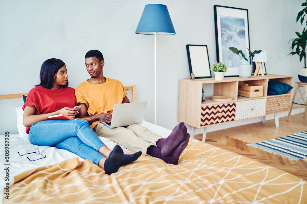 African american couple in love talking to each other sitting at bed room using laptop computer and smartphone for networking, dark skinned young family having conversation resting on free time.