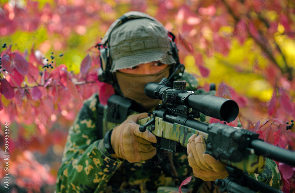 Closeup man in green uniform with sniper rifle. Airsoft soldier stand in red bush