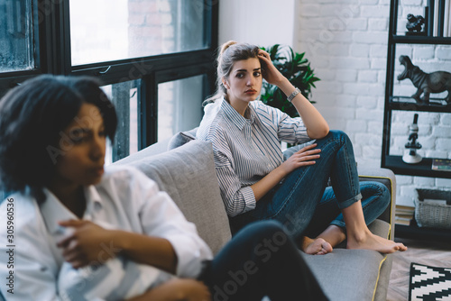 Worried caucasian young woman has conflict with offended african american hipster girl sitting on cozy couch at home.Unhappy multicultural friends don't talk after quarrel in modern apartment