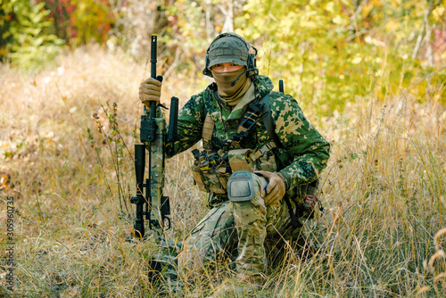 Airsoft man in uniform with sniper rifle, lurking in grass on forest background. Front view